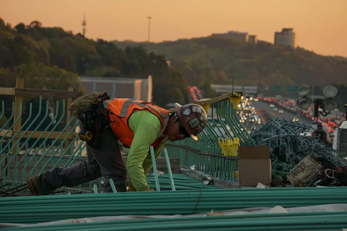 A construction worker in a hard hat and high-visibility vest working on rebar at dusk, highway traffic behind.