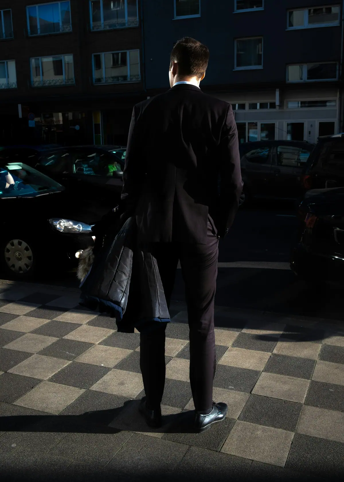 A man in a dark suit seen from behind, standing beside parked cars at night.